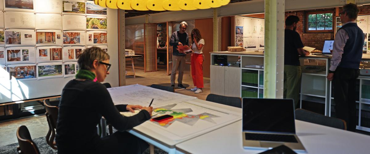 A busy architectural studio with a large, yellow, scalloped pendant light hanging over a table where a person is sketching, while colleagues collaborate in the background.