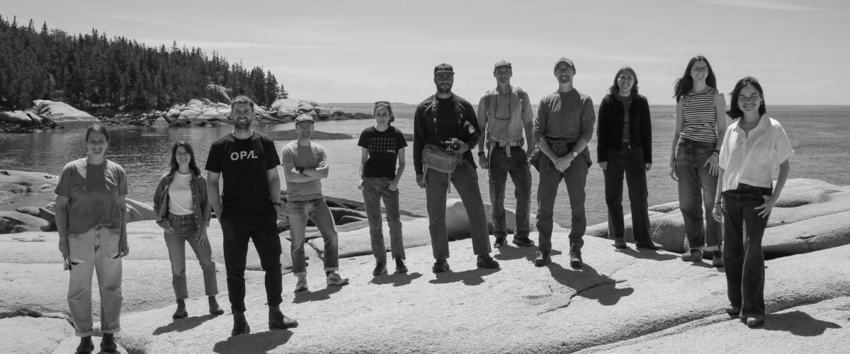 A black and white photo of the OPAL team standing across a rocky coastline with a view of the ocean behind.