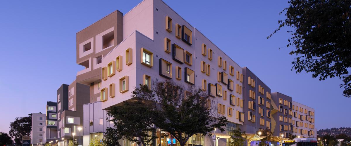 Exterior view of a multi-story student housing complex at dusk, featuring gray and white façades, projecting gold window surrounds, landscaped sidewalks, and a bus stop along the street.