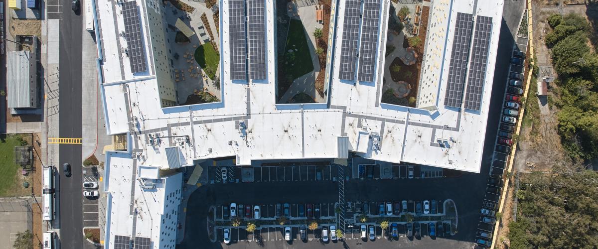Aerial view looking directly down on the student housing complex, showing its articulated building footprint, landscaped courtyards, rooftop solar panel arrays, white roofs, and adjacent parking.