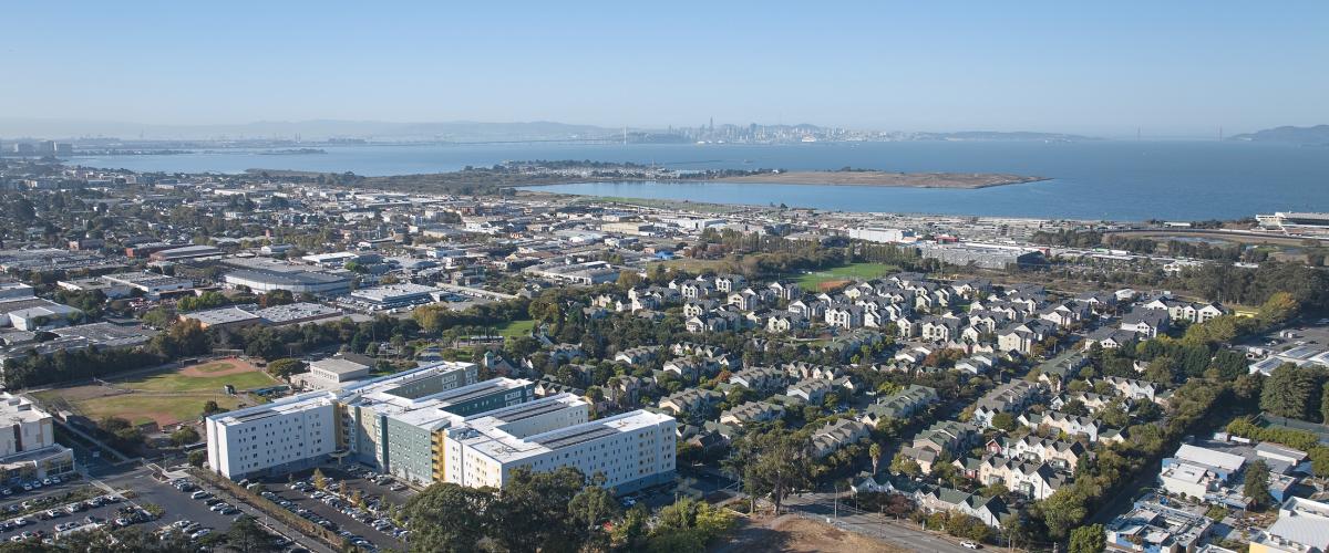 Wide aerial view of the student housing complex within its surrounding neighborhood, with residential streets, industrial buildings, San Francisco Bay, and the San Francisco skyline in the distance.