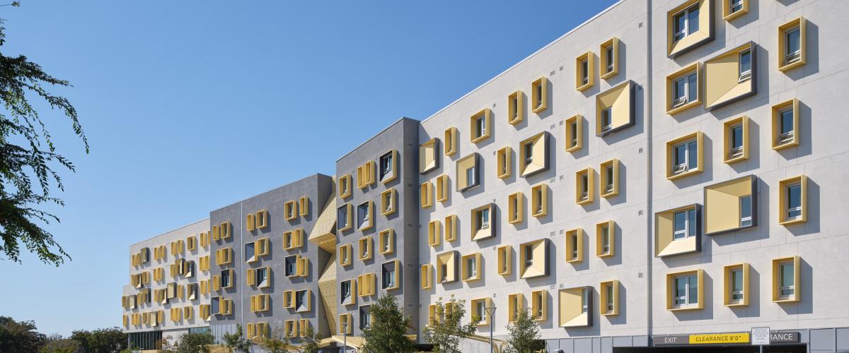 Street-level view of the long student housing façade, featuring gray and white exterior walls, projecting gold window surrounds, sculptural screened openings, landscaping, and a parking garage entrance.