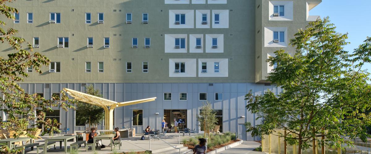 Landscaped residential courtyard with outdoor seating, pedestrian paths, shade structures, and trees, framed by a multi-story building with green-gray walls and recessed white window surrounds.