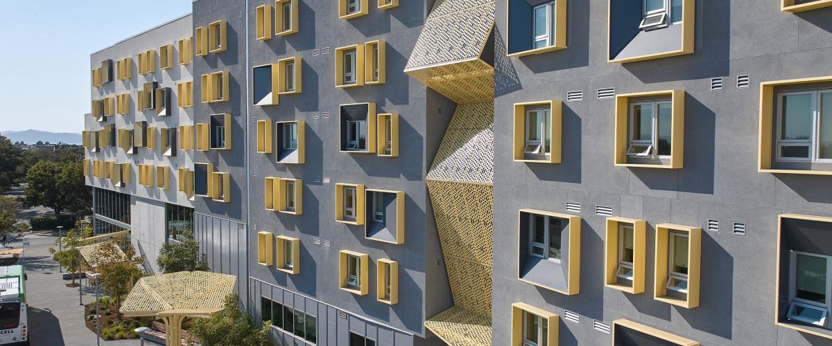 Close exterior view of the gray student housing façade with projecting gold window surrounds and sculptural perforated-metal screens shading outdoor spaces above landscaped sidewalks.