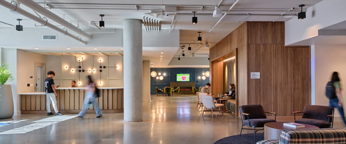 Open student housing lounge with polished concrete floors, exposed ceiling structure and utilities, wood-paneled walls, varied seating areas, and students gathering and moving through the space.