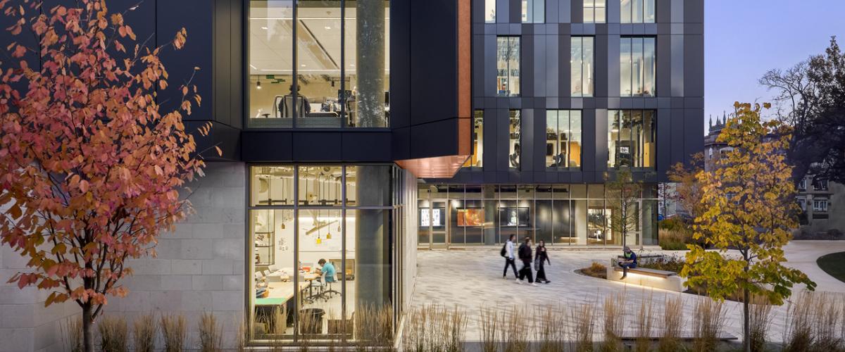 Exterior image of a modern academic building featuring a metal panel façade, large windows, illuminated entrance, landscaped planting beds, and pedestrians along the sidewalk.