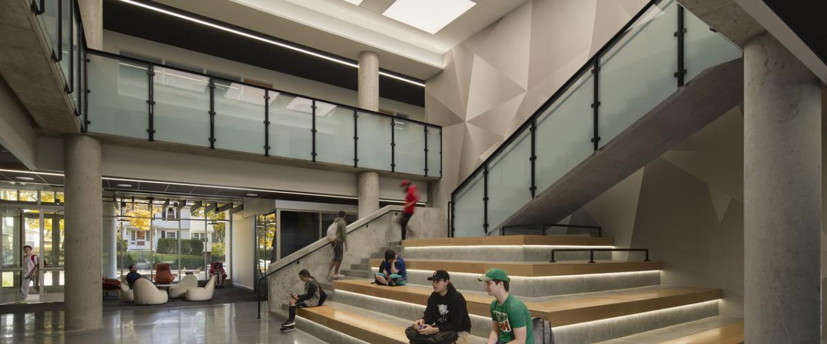 Students gather and study in a bright, multi-story campus commons featuring a tiered social stair, open circulation, glass railings, exposed concrete structure, and large ceiling light panels.