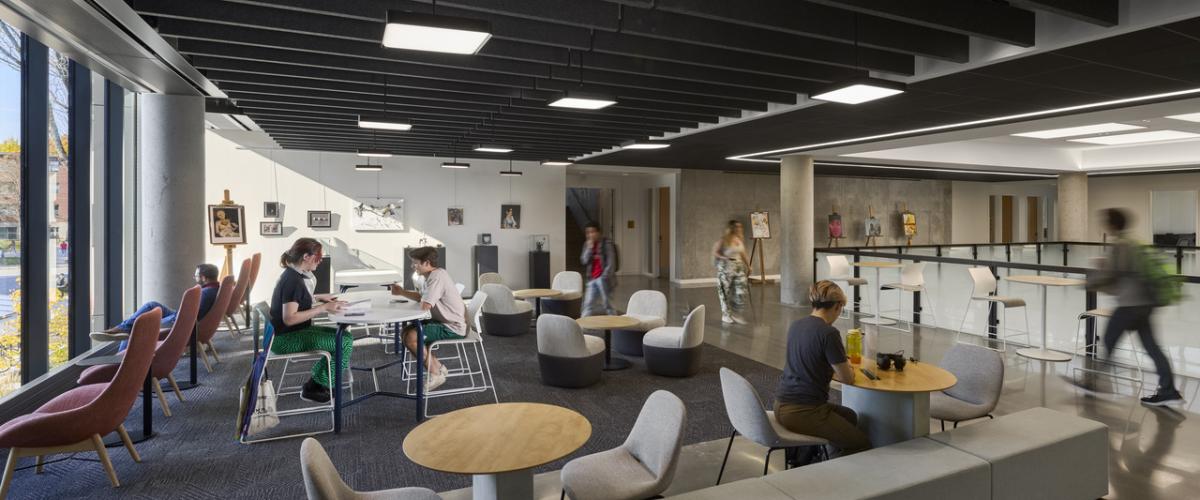 Students work individually and in groups throughout an open lounge with flexible seating, tables, artwork displays, and floor-to-ceiling windows providing daylight.
