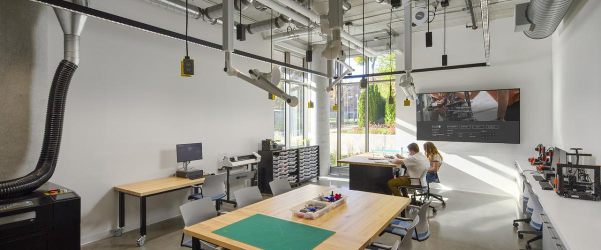 A makerspace equipped with digital fabrication tools, worktables, ventilation systems, and 3D printers supports hands-on learning while students collaborate at workstations.