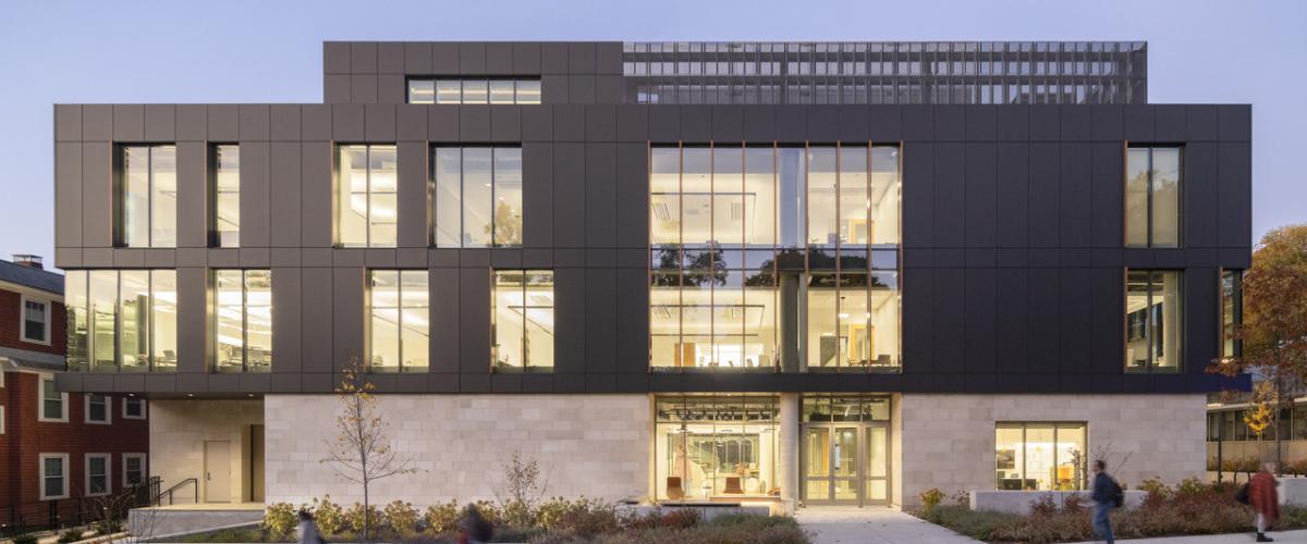 A contemporary academic building with a dark metal façade and expansive glazing frames a landscaped campus courtyard where students gather and walk between classes at dusk.