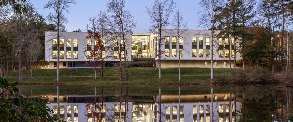 A contemporary white building situated on a grassy bank, surrounded by bare trees and reflected in a still lake.