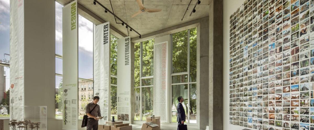 A modern, naturally lit exhibition space with large windows, concrete floors, and educational displays about building sustainability.