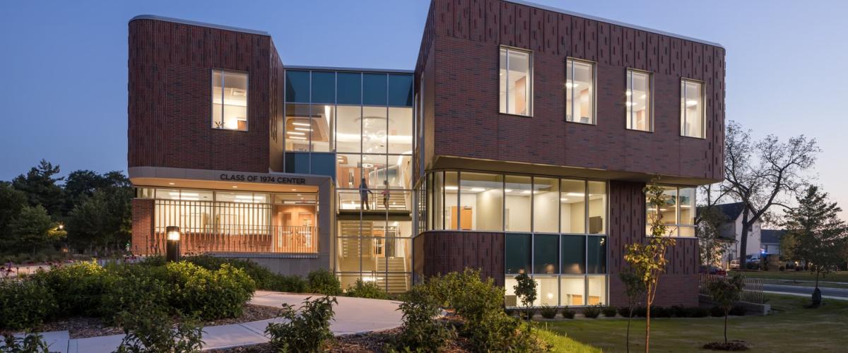 A modern, multi-story brick building with large glass windows, illuminated from within at dusk, featuring the "Class of 1974 Center."