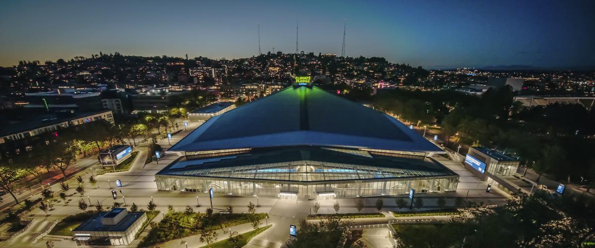 An aerial view of Climate Pledge Arena at dusk, featuring its distinctive, sweeping roofline and illuminated exterior.