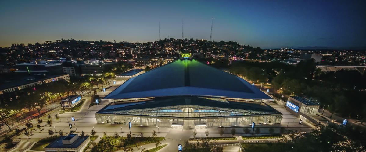 An aerial view of Climate Pledge Arena at dusk, featuring its distinctive, sweeping roofline and illuminated exterior.