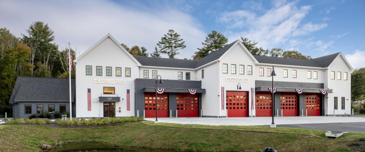 Exterior view of the station. A long white two story building with pitched New England style roof and features 6 large red garage doors