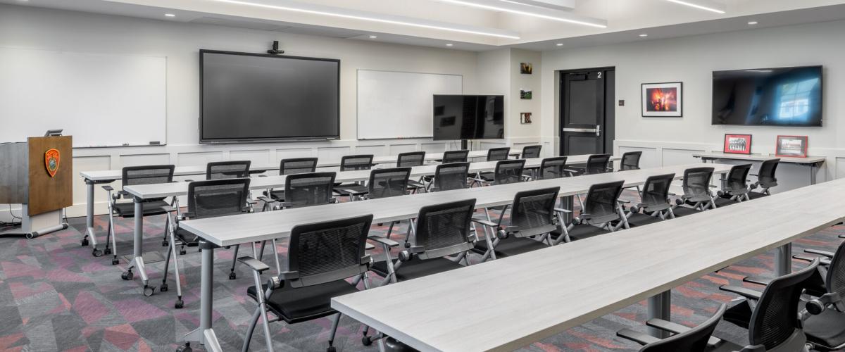 Classroom shot with four rows of long tables with chairs. A big tv with AV tech sits on the front wall with a lecture with a station emblem sits to one corner.