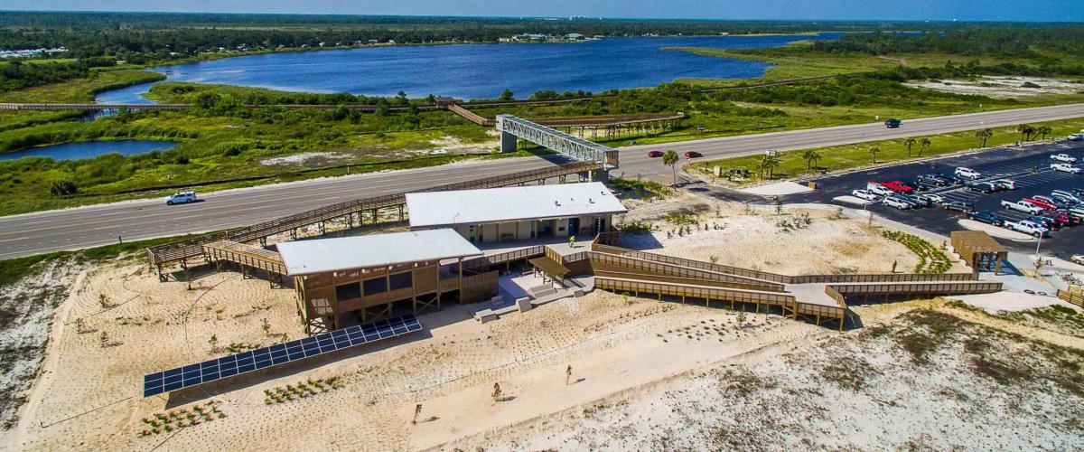 A coastal facility featuring two connected wooden structures with flat, white roofs. The building includes a covered porch area, large windows, and is flanked by a ground-mounted solar array on one side and extensive wooden boardwalk ramps on the other.