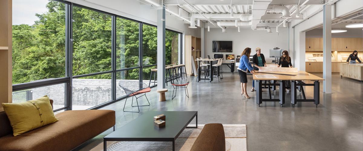 A bright, contemporary studio space with floor-to-ceiling windows, modern furniture, and a group of people working together at a large central table.