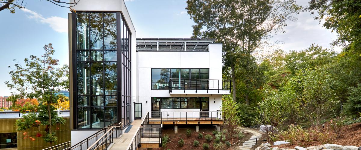 An exterior view of a contemporary, multi-level white building surrounded by trees, with a prominent glass stairwell and a wooden ramped walkway.