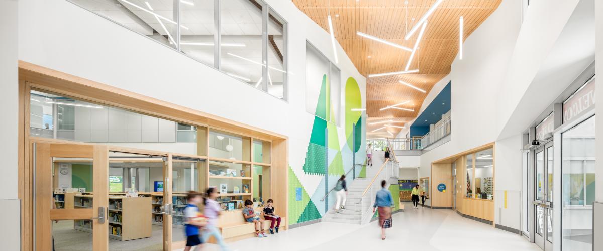 The sunny lobby and main entrance of Hardy Elementary School. The space features wood detailing and ceiling panels, nature themed environmental graphics, and a central staircase. Young students move about the space.