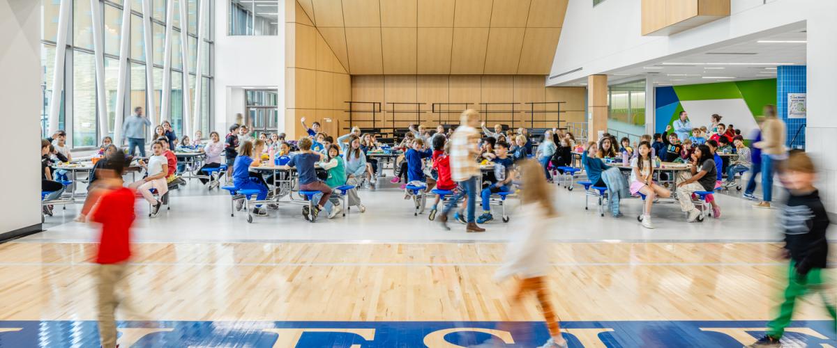 View of a bustling cafeteria seen from an adjacent gymnasium, with students eating at tables near floor‑to‑ceiling windows.