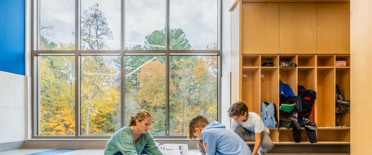 Students and teacher sitting on the floor near large windows, working on a project beside wooden cubbies.