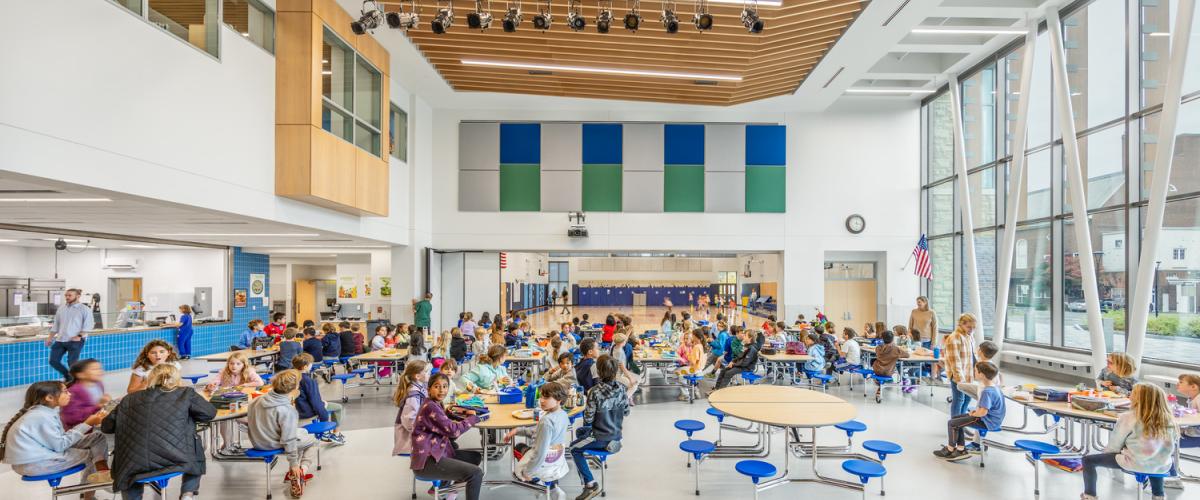 Large, bright cafeteria with students seated at blue tables under a wood‑slatted ceiling and tall glass windows.
