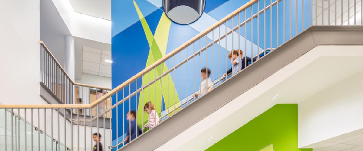Open multi‑level school common area with a brightly painted staircase, geometric lighting, and students sitting and walking throughout the space.