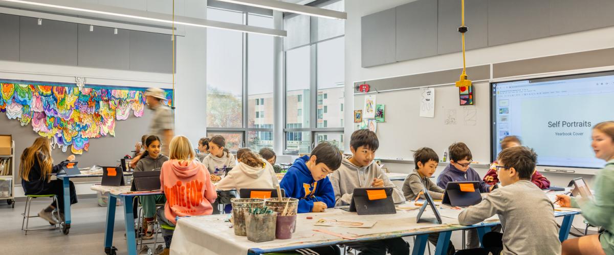 Art room with students working at tables using tablets and art supplies, with colorful artwork displayed on the wall.