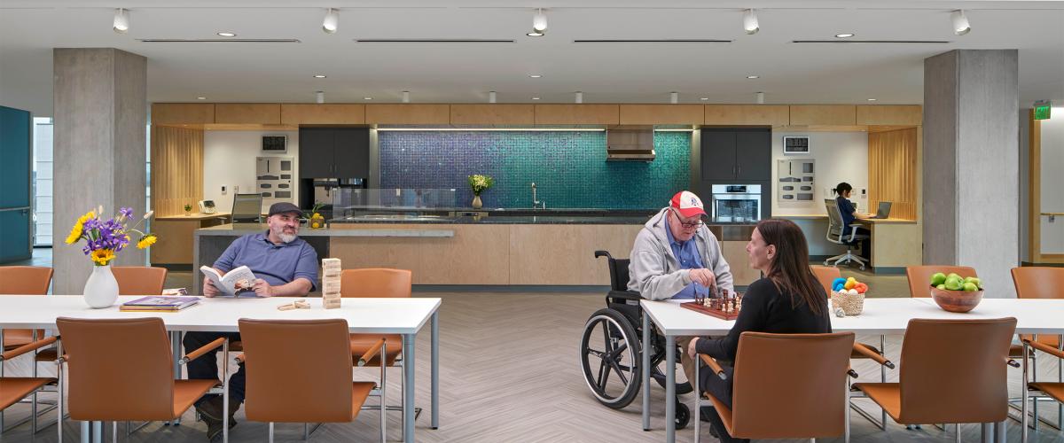 A shared kitchen space. Some residents at sitting at communal tables featuring a game of chess and a game of Jenga.