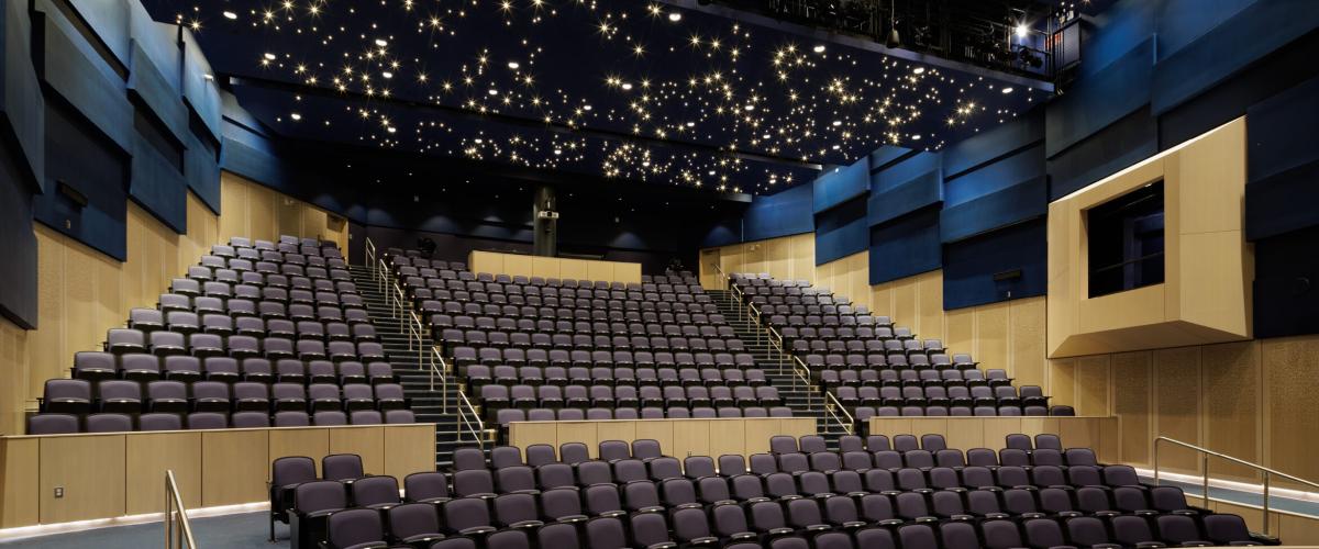 The view of the schools auditorium looking from the stage to the seating. The photo highlights the room's velvety deep blue wall panels and a ceiling with a light installation that resembles stars in the night sky