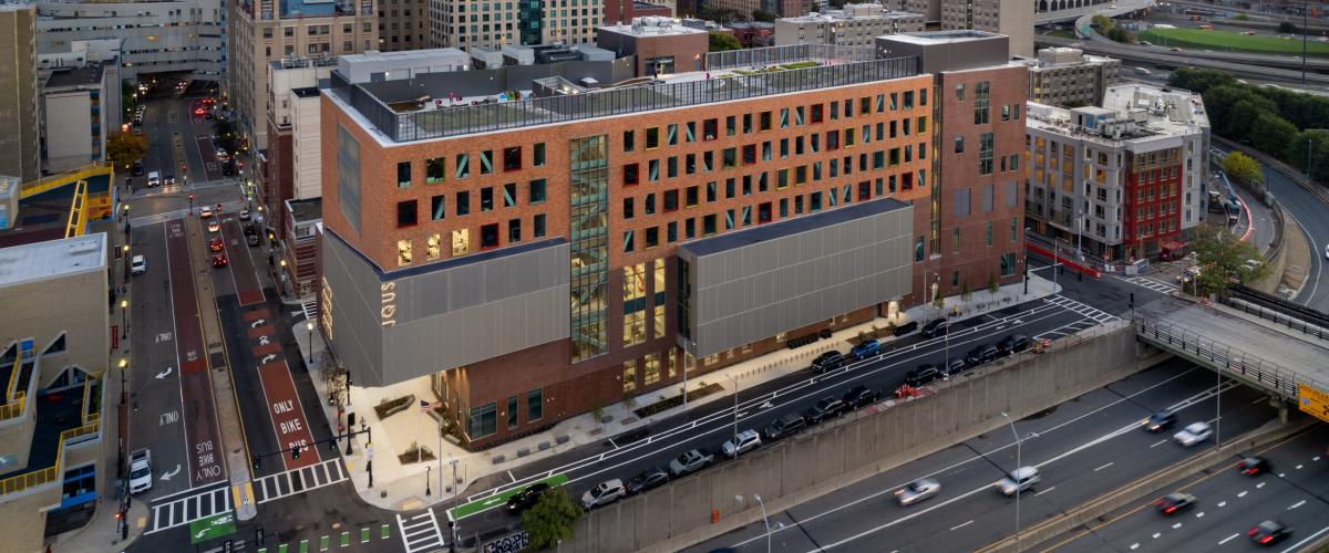 An aerial photo of the school. A seven story building with a rooftop green space, two silver volumes of building sticking our to the left and middle, and a two tone brick facade featuring windows with irregular protruding frame with bold colors of red, yellow, green, and blue. The school occupies a narrow strip of a city block that sits adjacent to the visible subterranean highway.