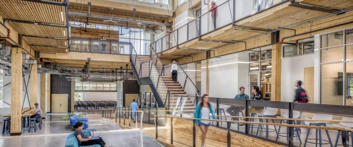 A spacious, modern building interior featuring exposed wooden ceilings, a central staircase, and people working in various lounge and desk areas.
