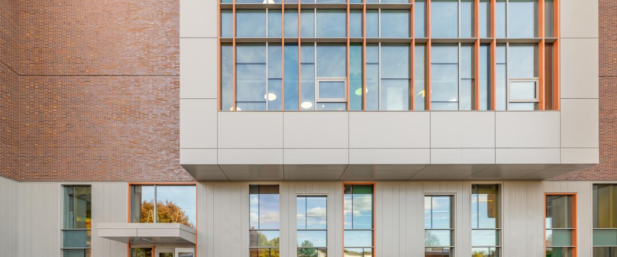 The back entrance of Henry K. Oliver Elementary & Middle Schools, featuring a modern façade with large vertical windows and brick and metal paneling. A sign with the school’s name is in front of the entry walkway, and students are walking in and out of the building.