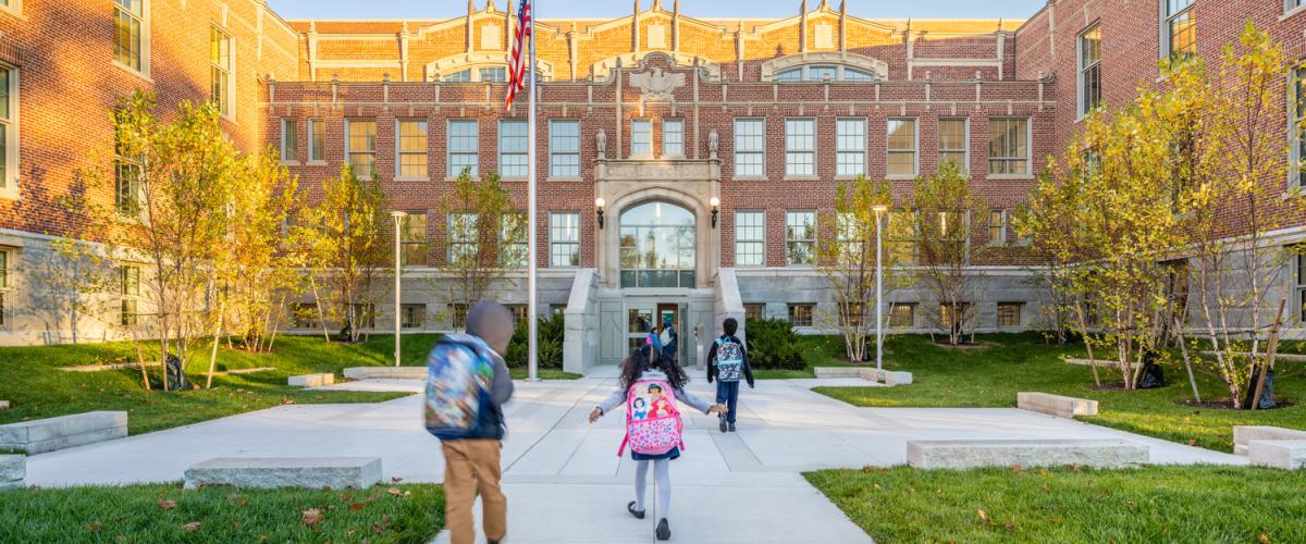 A historic brick school building with large windows and an ornate central entrance. A flagpole stands in front of a landscaped courtyard with walkways leading to the doors. Students with backpacks are walking toward the entrance.