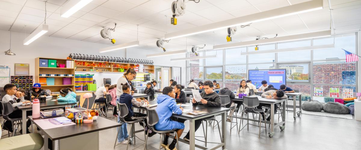 A brightly lit classroom with large windows and multiple tables where students are seated and working on assignments. The room has open shelving filled with colorful supplies along the back wall and ceiling-mounted equipment. A teacher stands near one of the tables assisting students.