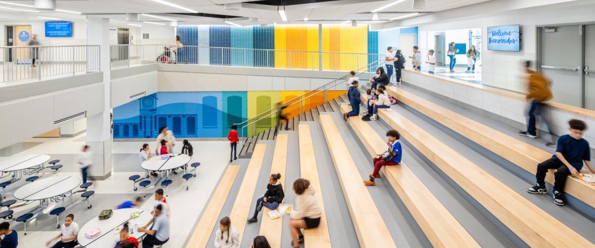 A large, open school common area with wide tiered wooden seating on one side and round cafeteria tables on the lower level. Students are seated on the steps and walking through the space. A multicolored wall mural and geometric art panels decorate the background.