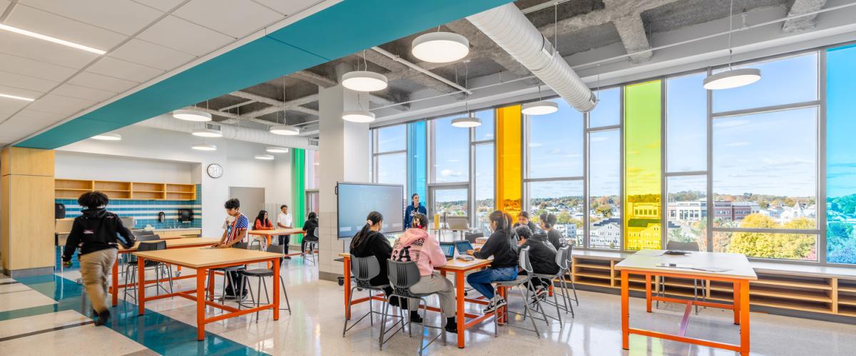 A spacious breakout area with tall windows featuring brightly colored vertical panels. Several tables are arranged throughout the room, and students are seated while an instructor stands near a smartboard at the front. The room has teal and white flooring and exposed ceiling elements.