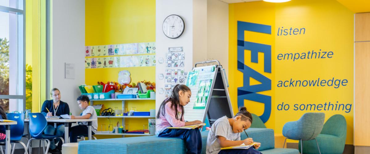 A colorful student common area with bright yellow walls, teal seating, and children reading or working on tablets. Artwork is displayed on the wall, and an inspirational message painted vertically reads ‘listen, empathize, acknowledge, do something.’ A small group workspace is visible in the background.