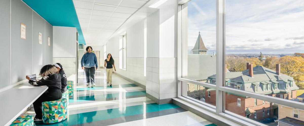 A school hallway with turquoise and white patterned flooring and large windows overlooking nearby buildings. A built-in counter with patterned stools provides a study area where a student is seated, while others walk down the hall.