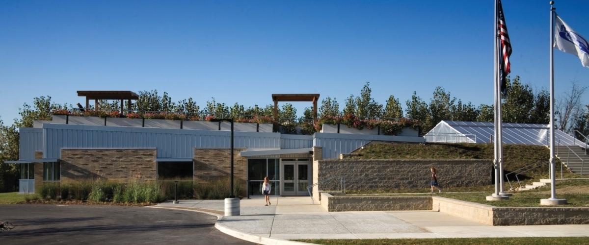 A low-profile, modern institutional building with a flat roof supporting a green roof, a central glass entrance, and an adjacent greenhouse structure.