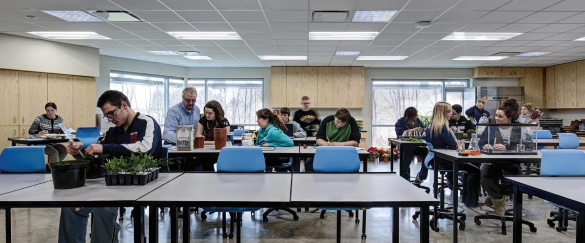 Students in a simple classroom interior with polished concrete floors, modular lab tables, and large windows providing natural light.