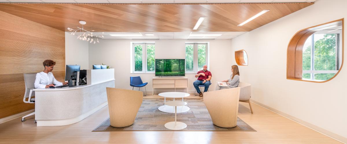 Photo of a well-lit healthcare center lobby with people seated in the waiting area and behind a check-in desk. Interior materials consist of light woods and organic forms that prioritize ample natural light.