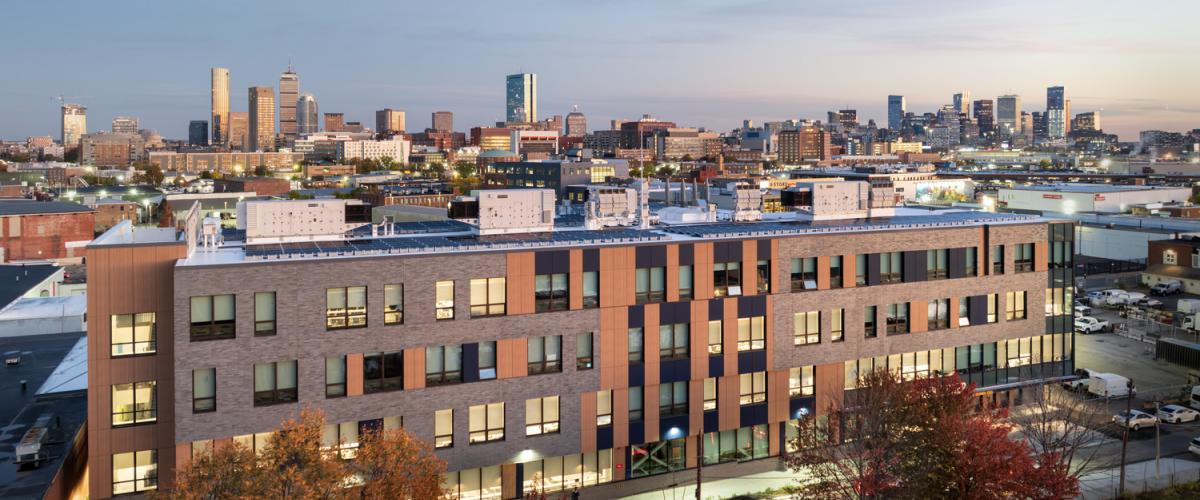Four‑story Roxbury Prep building at dusk with the Boston skyline in the background and fall trees in the foreground.