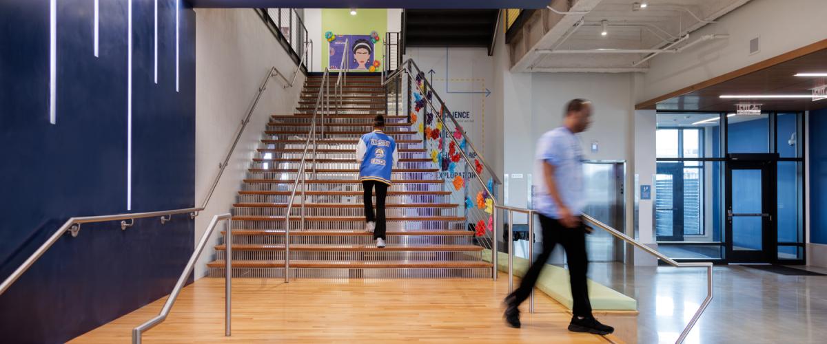 Modern school lobby with a wide wooden stair landing leading to a central staircase, with colorful student artwork and a mural visible on the upper level.