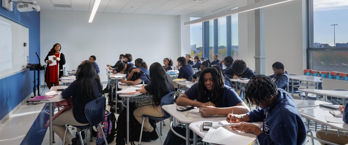 A bright classroom with large windows where students sit at grouped desks while a teacher stands at the whiteboard.