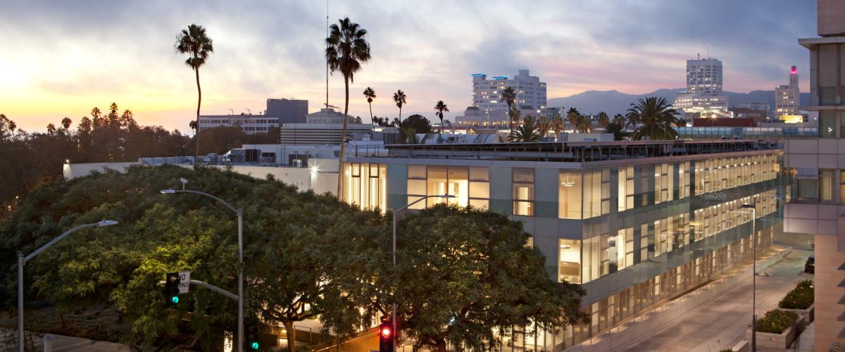 A contemporary multi-story building with large windows, glowing from within against a twilight sky. Palm trees and streetlights are visible in the foreground, with a distant city skyline and mountains behind.