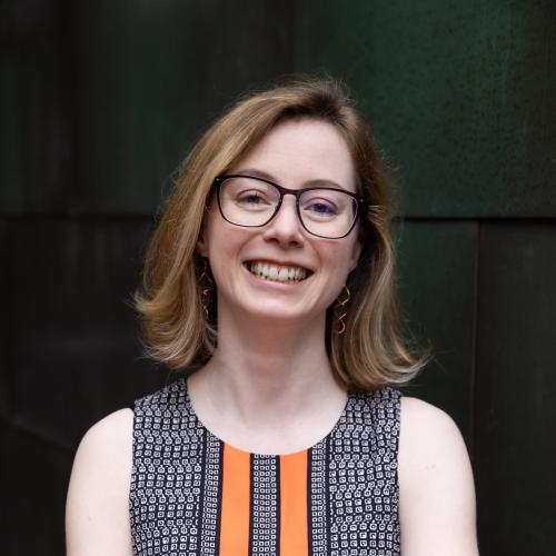portrait headshot of a young professional woman with glasses and honey colored shoulder length hair. she is wearing a patterned dress with orange stripes