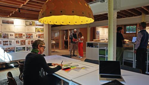 A busy architectural studio with a large, yellow, scalloped pendant light hanging over a table where a person is sketching, while colleagues collaborate in the background.